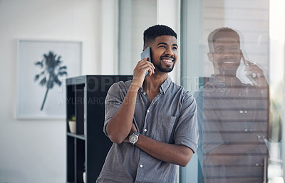 Buy stock photo Shot of a young businessman talking on a cellphone while standing at a window in an office