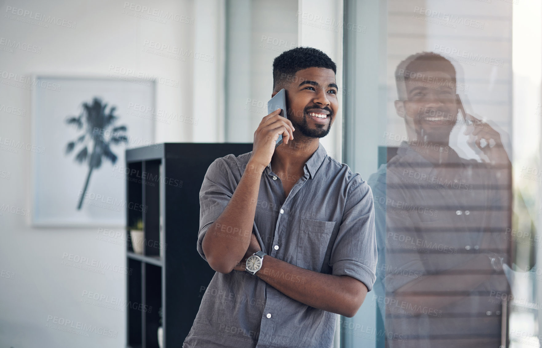 Buy stock photo Shot of a young businessman talking on a cellphone while standing at a window in an office