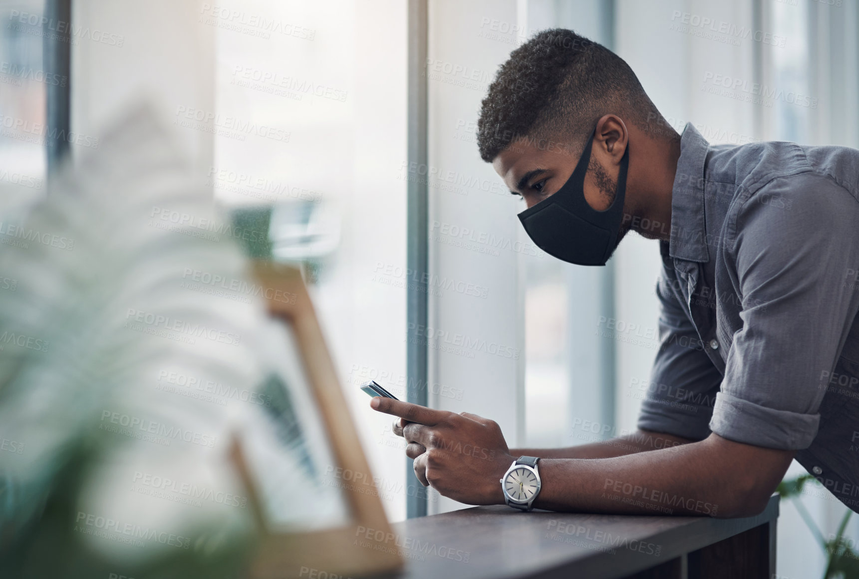 Buy stock photo Shot of a young businessman wearing a face mask while using a cellphone in an office
