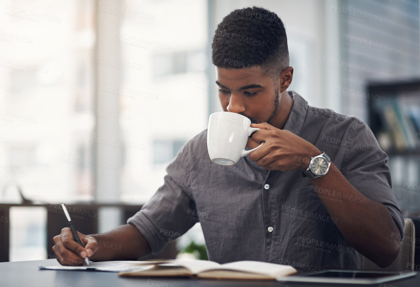 Buy stock photo Shot of a young businessman drinking coffee while going through paperwork in an office