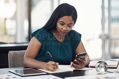 Buy stock photo Shot of a young businesswoman writing notes while using a cellphone in an office