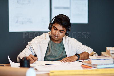 Buy stock photo Shot of a young architect wearing headphones and writing in a notebook at his desk in a modern office
