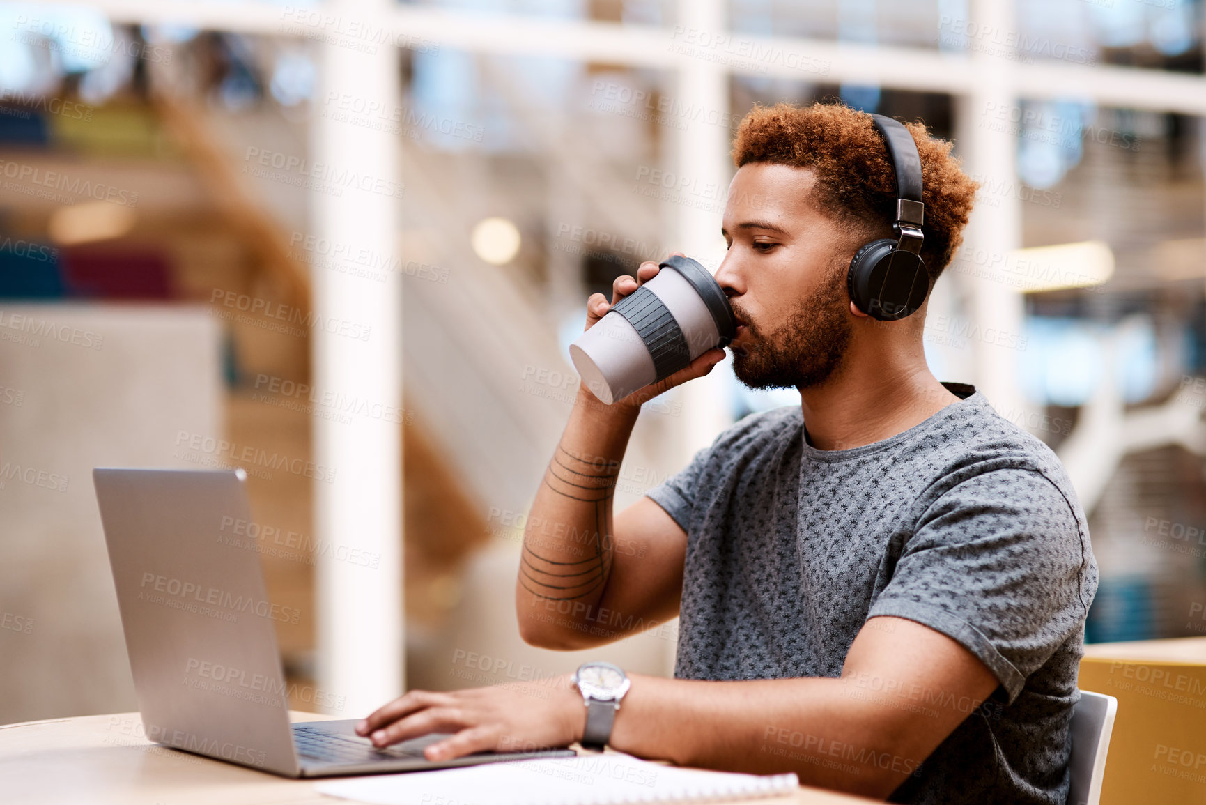 Buy stock photo Shot of a young businessman wearing headphones and drinking coffee while working on a laptop in an office