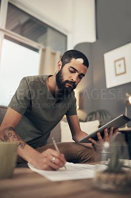 Buy stock photo Shot of a young man using a digital tablet while going through paperwork at home