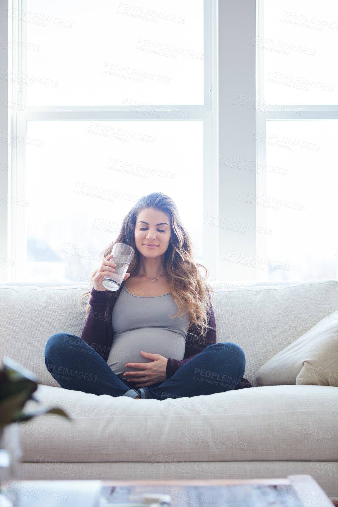 Buy stock photo Full length shot of an attractive young pregnant woman drinking water while sitting on the sofa at home