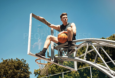 Buy stock photo Shot of a sporty young man sitting on a basketball hoop on a sports court