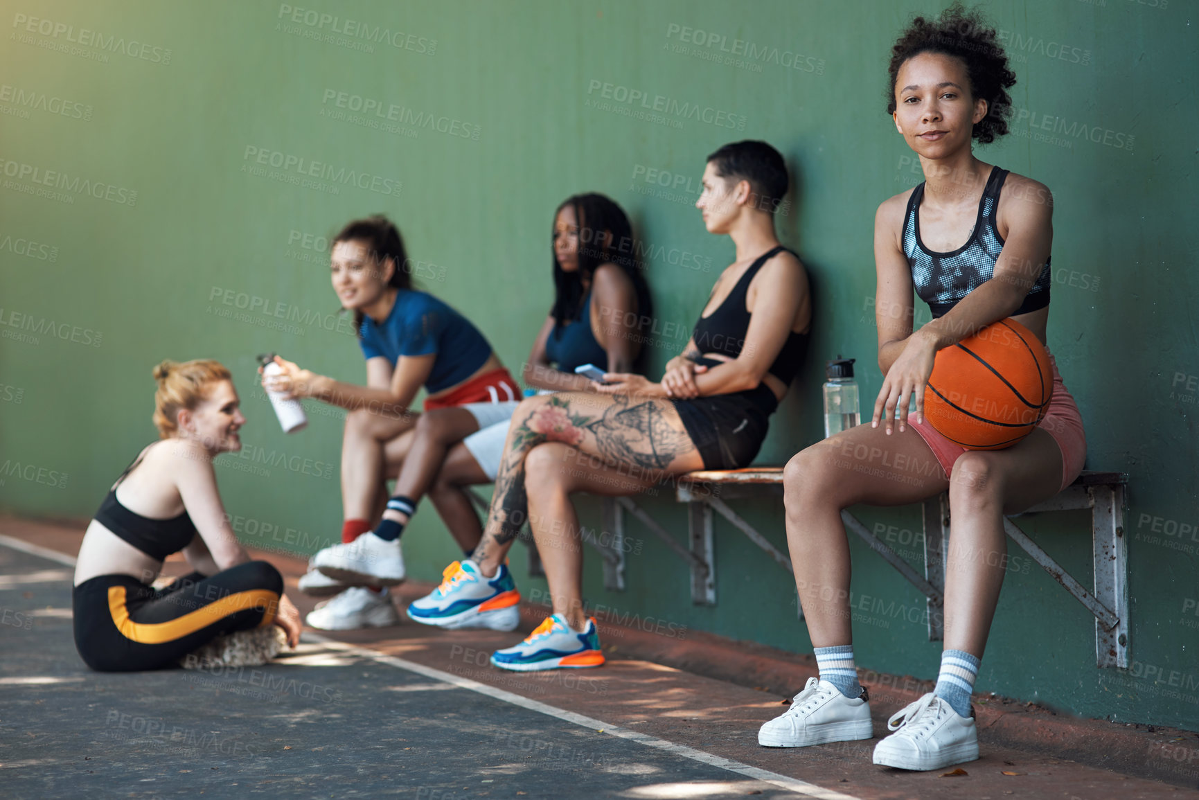 Buy stock photo Full length portrait of an attractive young female athlete sitting on a bench at the basketball court with her teammates in the background