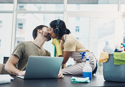 Buy stock photo Shot of a young woman kissing her husband while she cleans the house and he uses a laptop