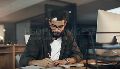 Buy stock photo Shot of a young businessman writing in a notebook and using a computer during a late night in a modern office