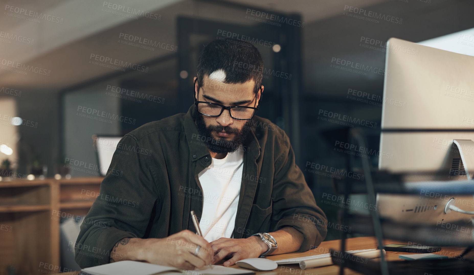 Buy stock photo Shot of a young businessman writing in a notebook and using a computer during a late night in a modern office