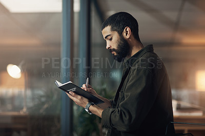 Buy stock photo Shot of a young businessman writing in a notebook during a late night in a modern office