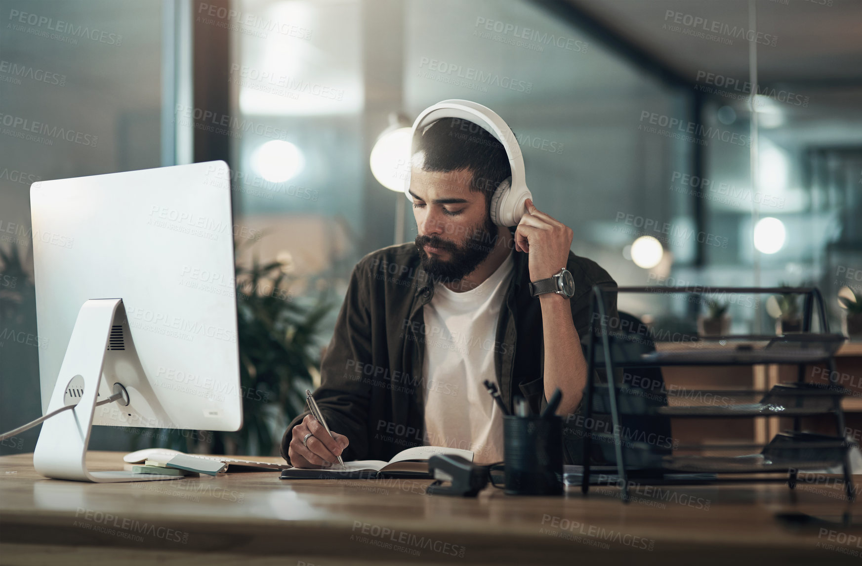 Buy stock photo Shot of a young businessman writing in a notebook and using a computer with headphones during a late night at work