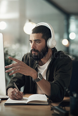 Buy stock photo Shot of a young businessman writing in a notebook and using a computer with headphones during a late night at work