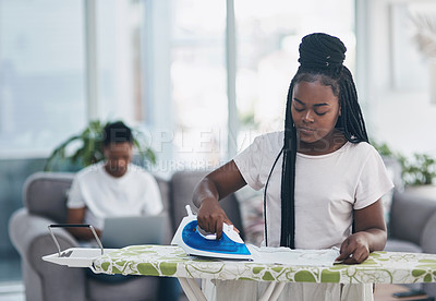 Buy stock photo Shot of a young woman ironing clothing with her husband on the sofa in the background