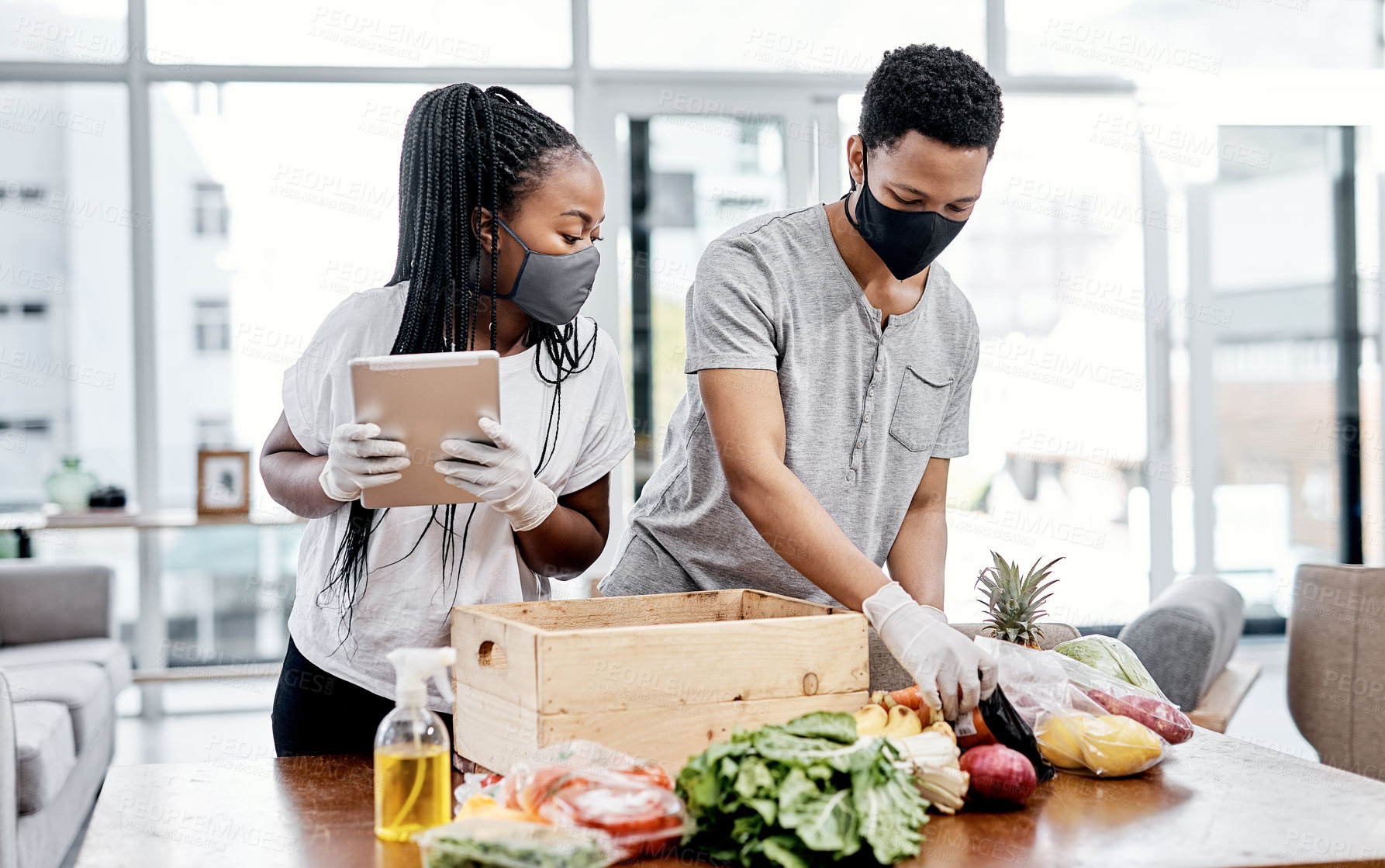 Buy stock photo Shot of a masked young couple using a digital tablet while disinfecting their groceries at home