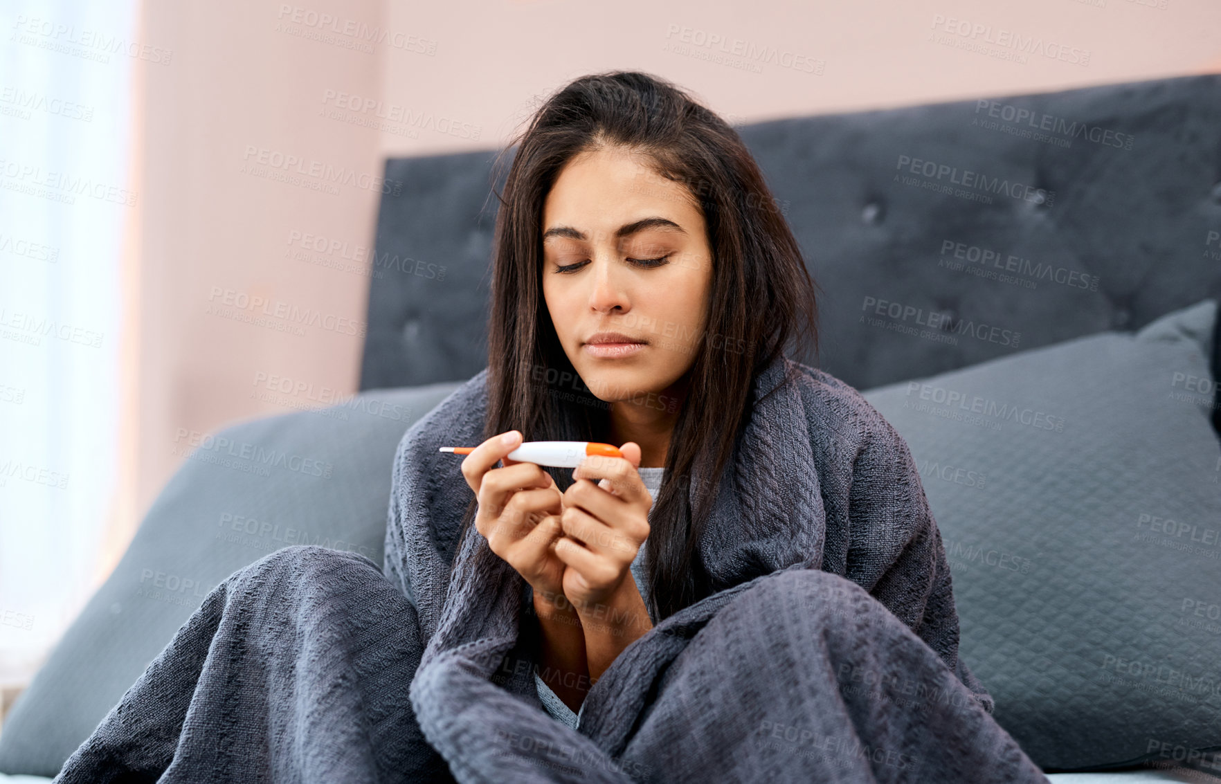 Buy stock photo Shot of a young woman taking her temperature while recovering from an illness in bed at home