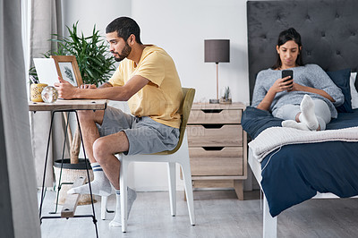 Buy stock photo Shot of a young man working from home while his wife uses a smartphone on the bed