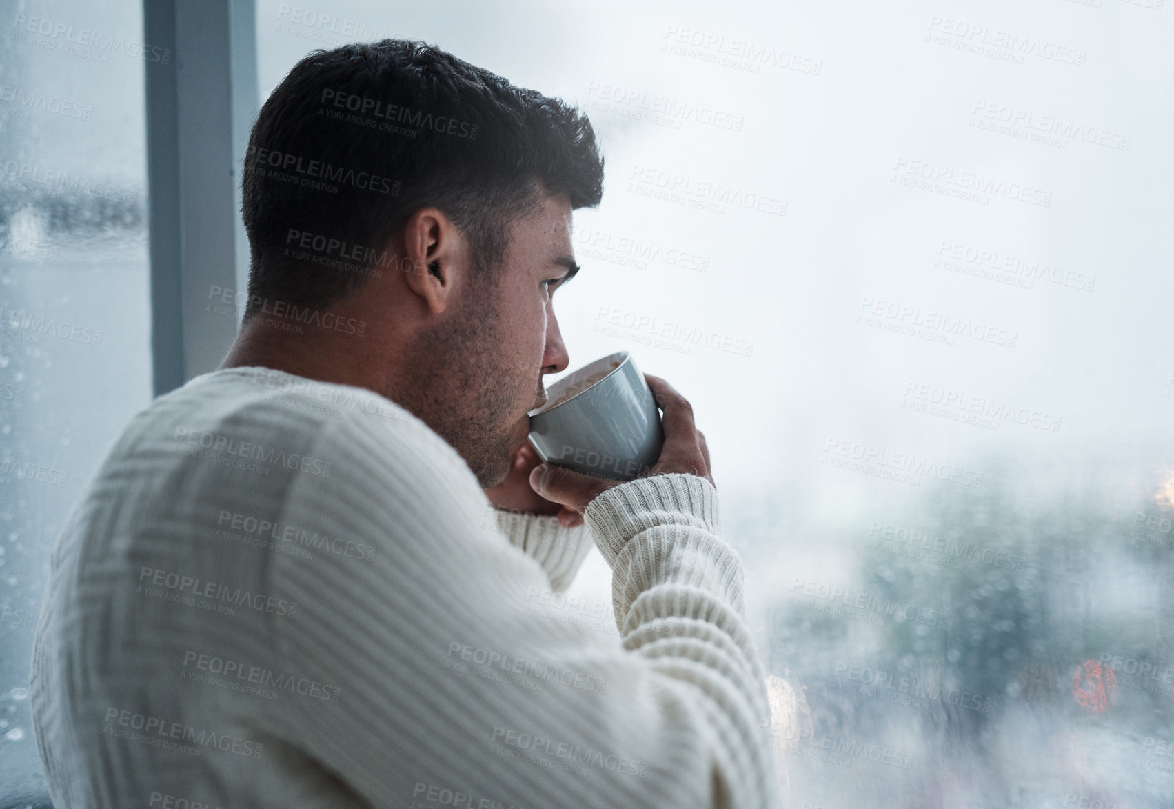 Buy stock photo Shot of a young man having coffee and looking out of a window on a rainy day at home