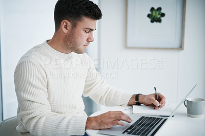 Buy stock photo Shot of a young man using a laptop and writing in a notebook while working from home