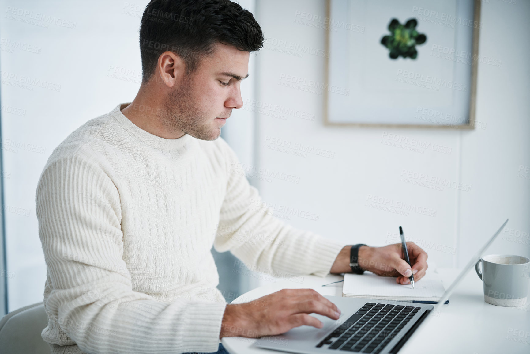 Buy stock photo Shot of a young man using a laptop and writing in a notebook while working from home