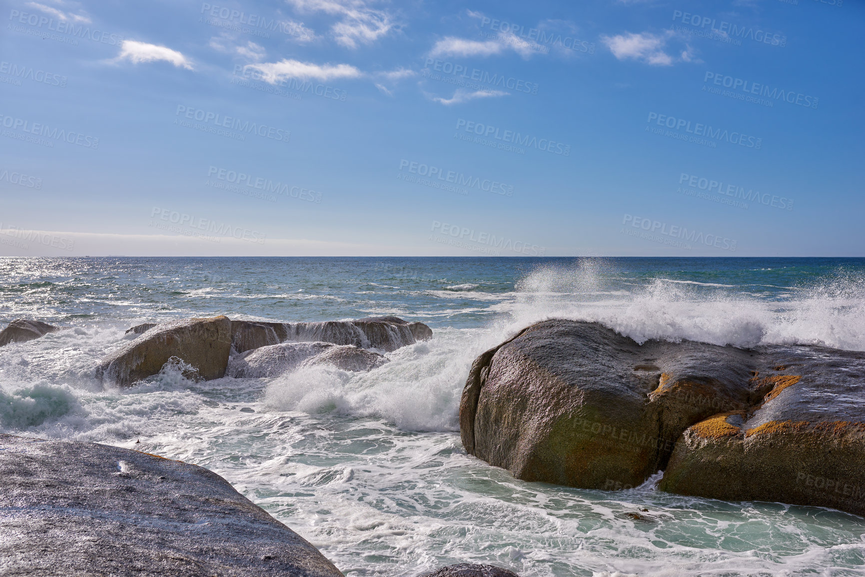 Buy stock photo Beautiful landscape view of the rough ocean waves breaking on large boulders with a blue sky background. Tidal waves crashing into rocks on the windy wild coast and beach of Cape Town, South Africa