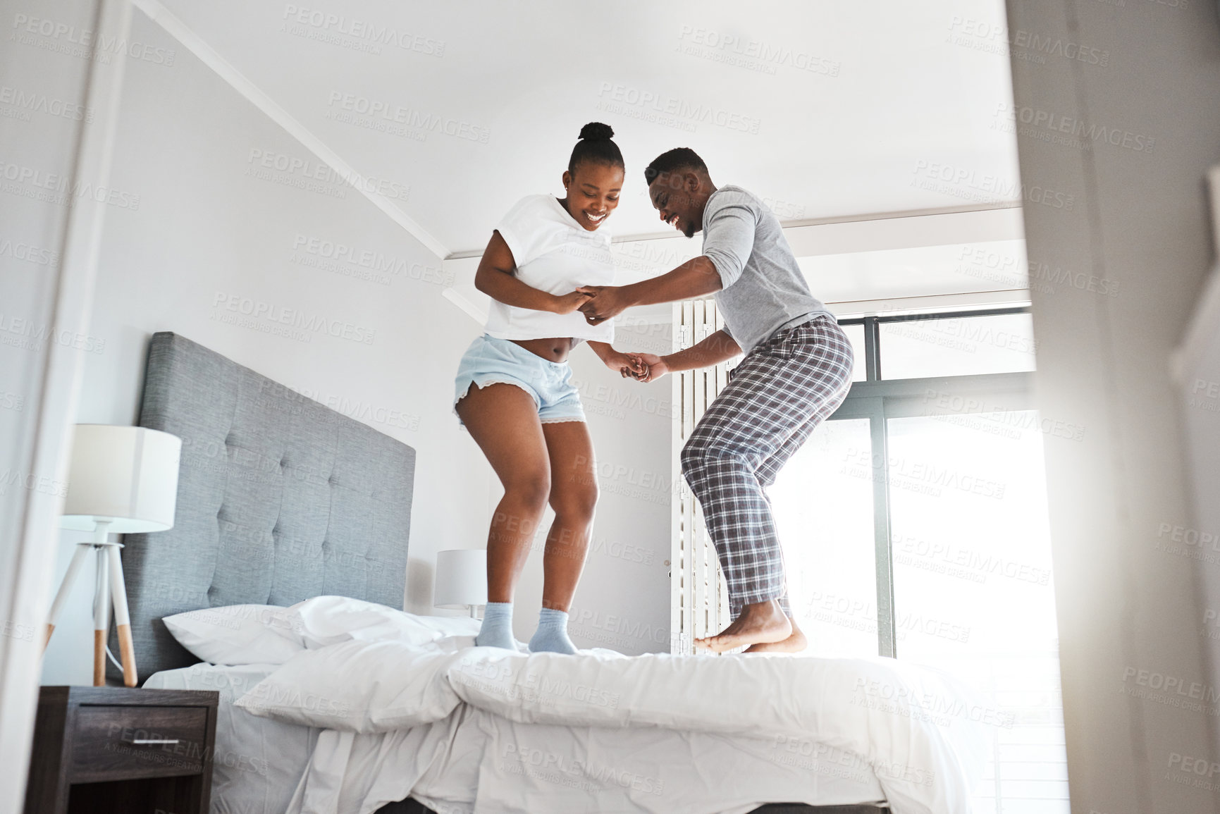 Buy stock photo Shot of a young couple jumping playfully together on a bed at home