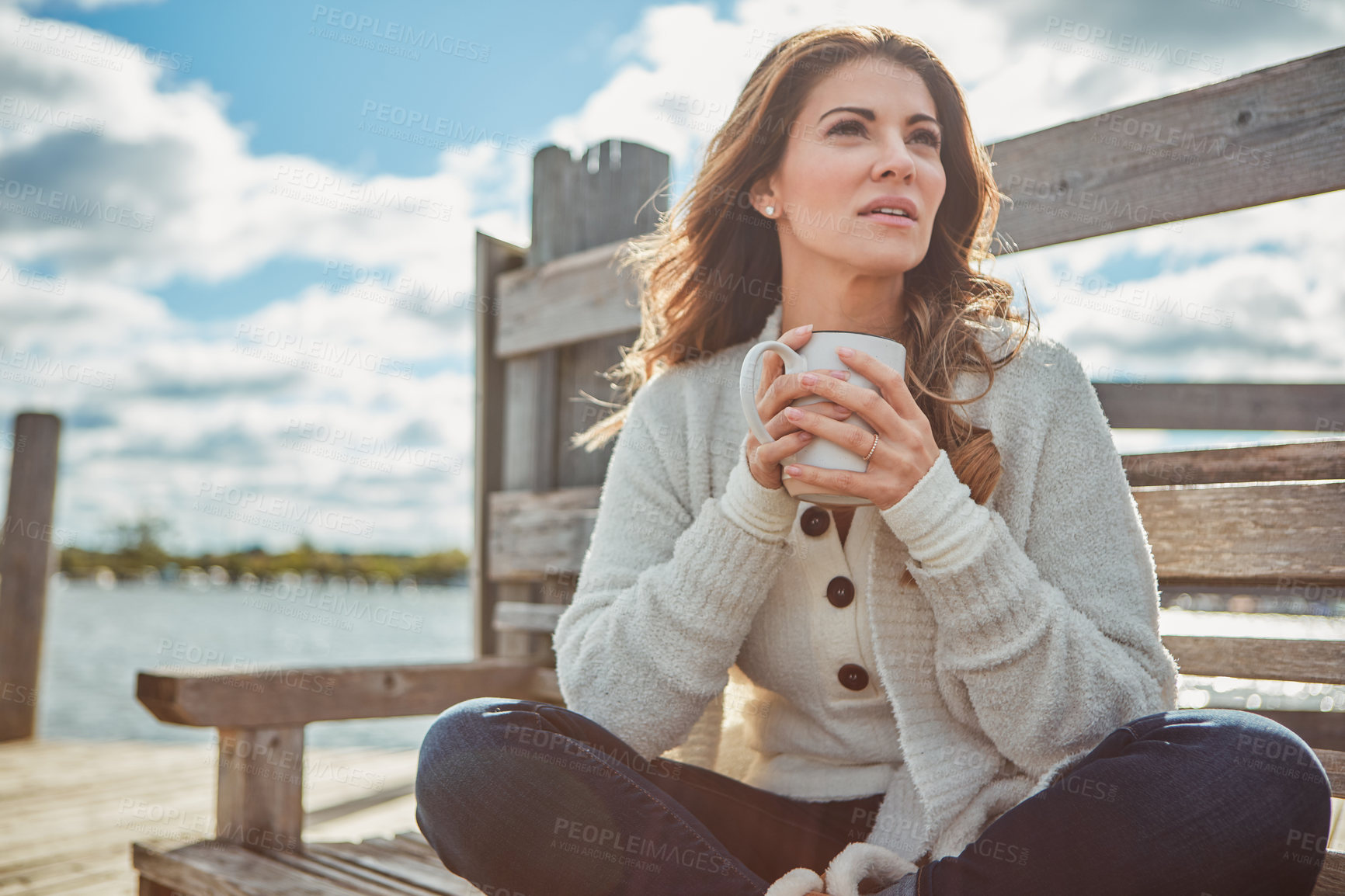 Buy stock photo Shot of a beautiful young woman enjoying a warm beverage while relaxing on a bench at a lake