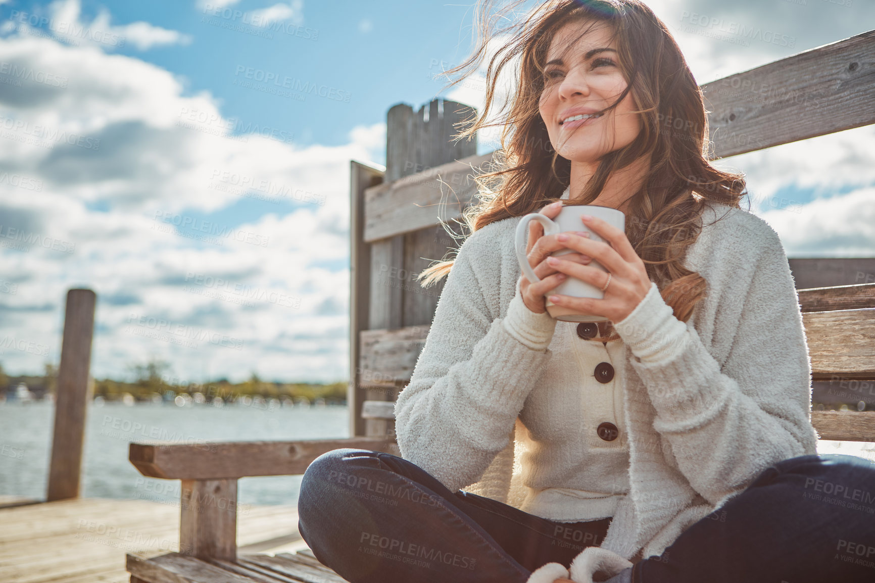 Buy stock photo Shot of a beautiful young woman enjoying a warm beverage while relaxing on a bench at a lake