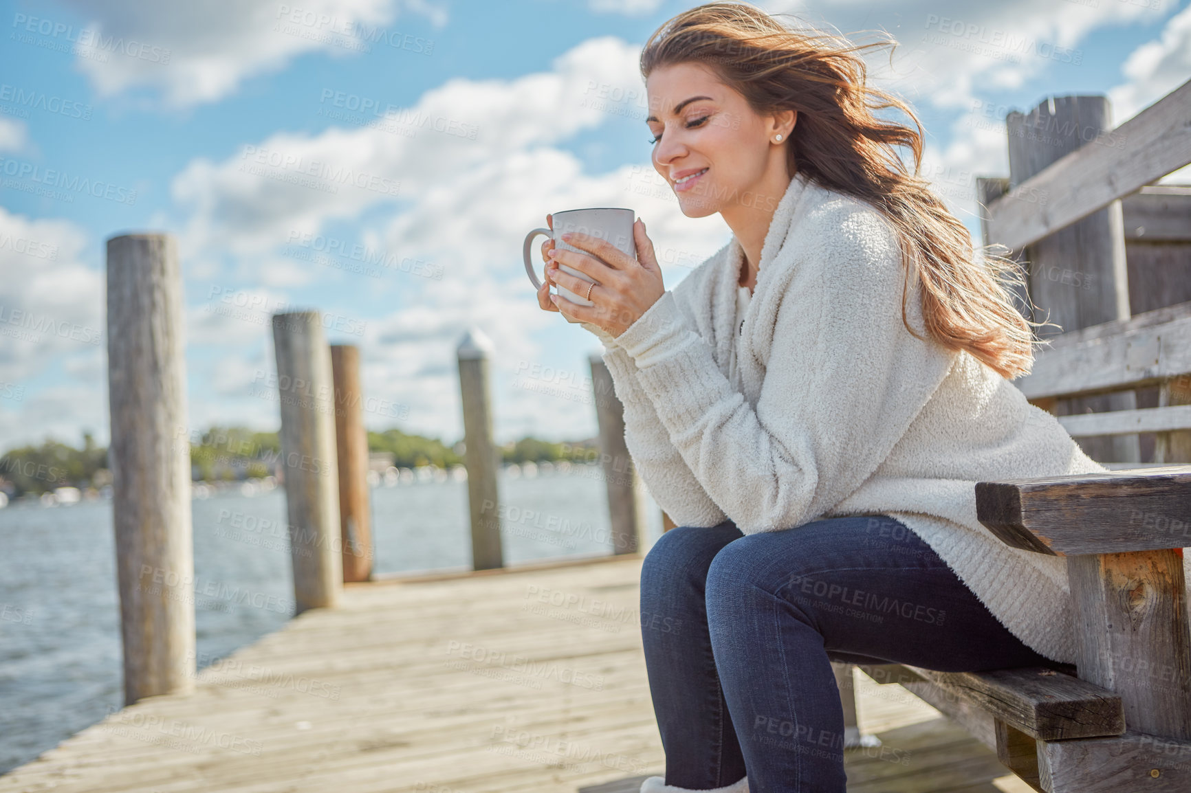 Buy stock photo Shot of a beautiful young woman enjoying a warm beverage while relaxing on a bench at a lake