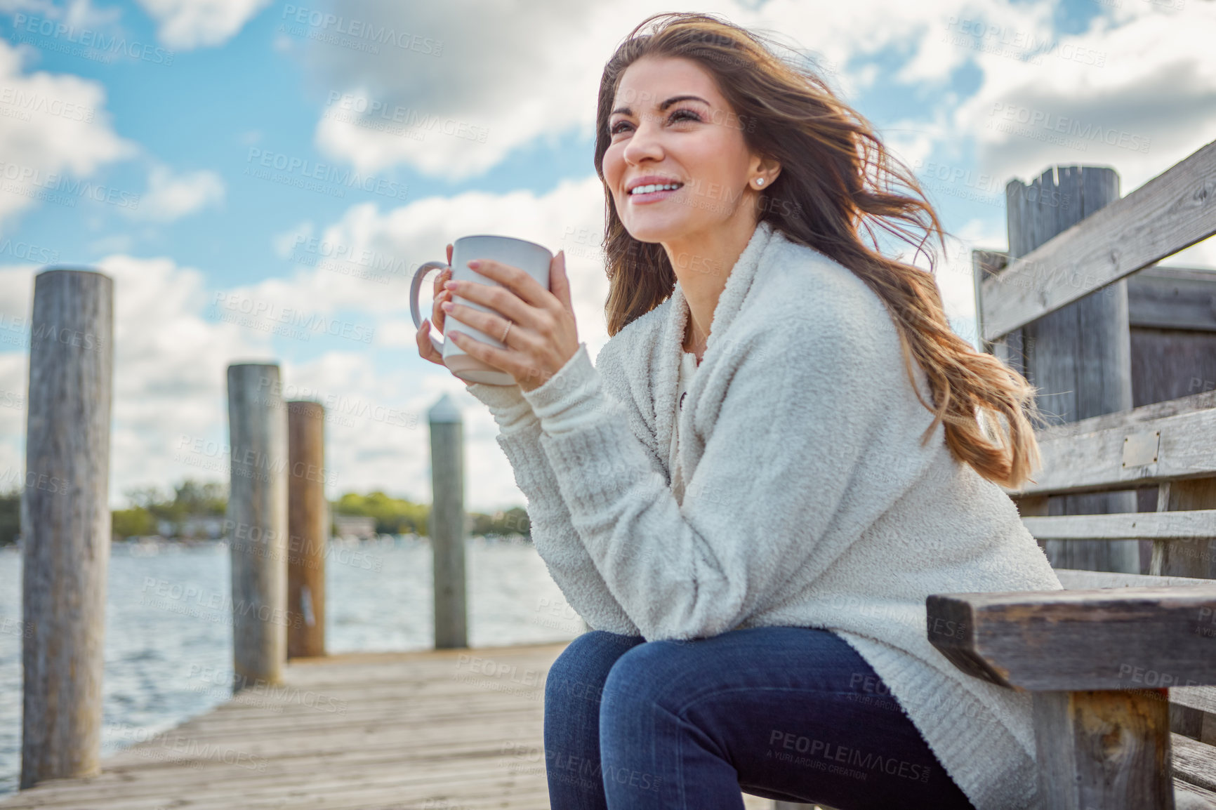 Buy stock photo Shot of a beautiful young woman enjoying a warm beverage while relaxing on a bench at a lake