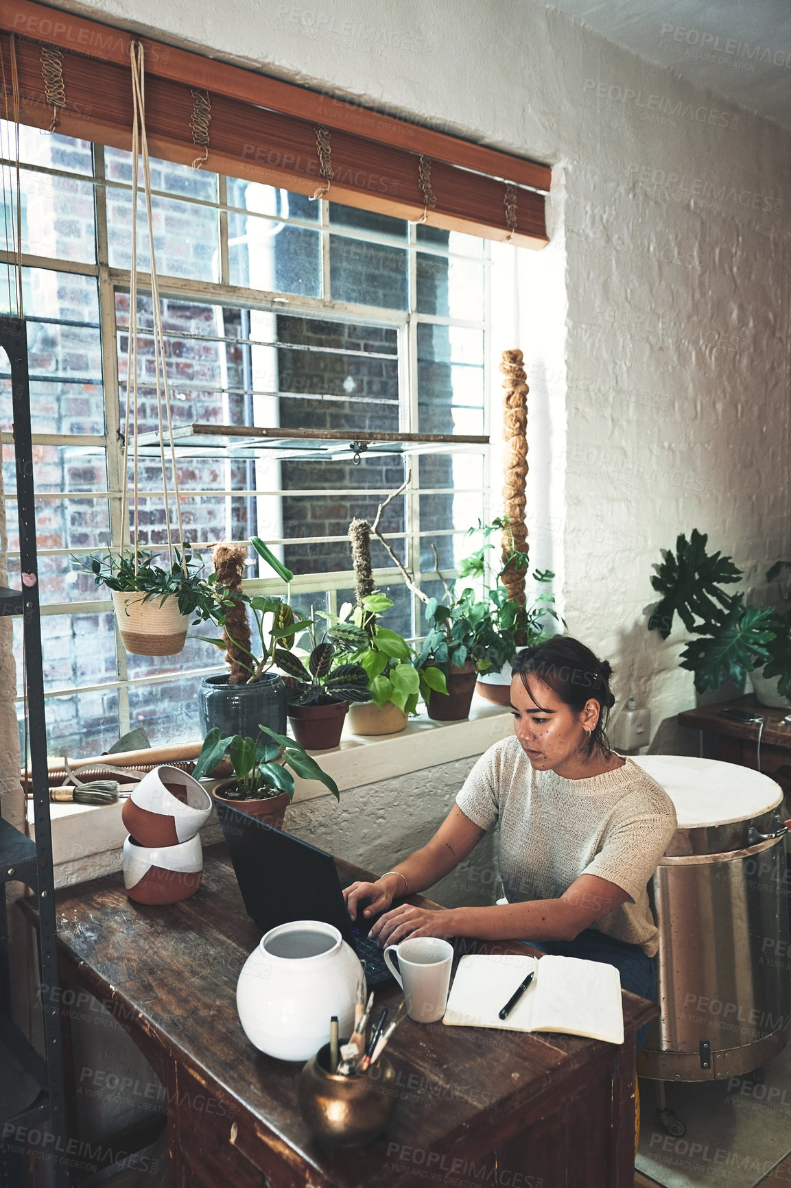 Buy stock photo Cropped shot of an attractive young business owner sitting alone in her pottery studio and using her laptop