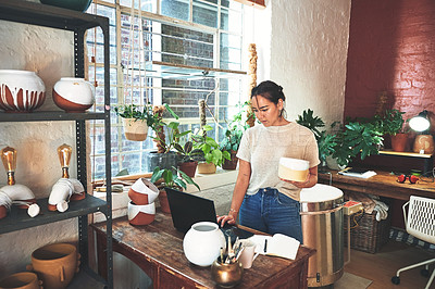 Buy stock photo Cropped shot of an attractive young business owner standing and holding a clay pot while using her laptop