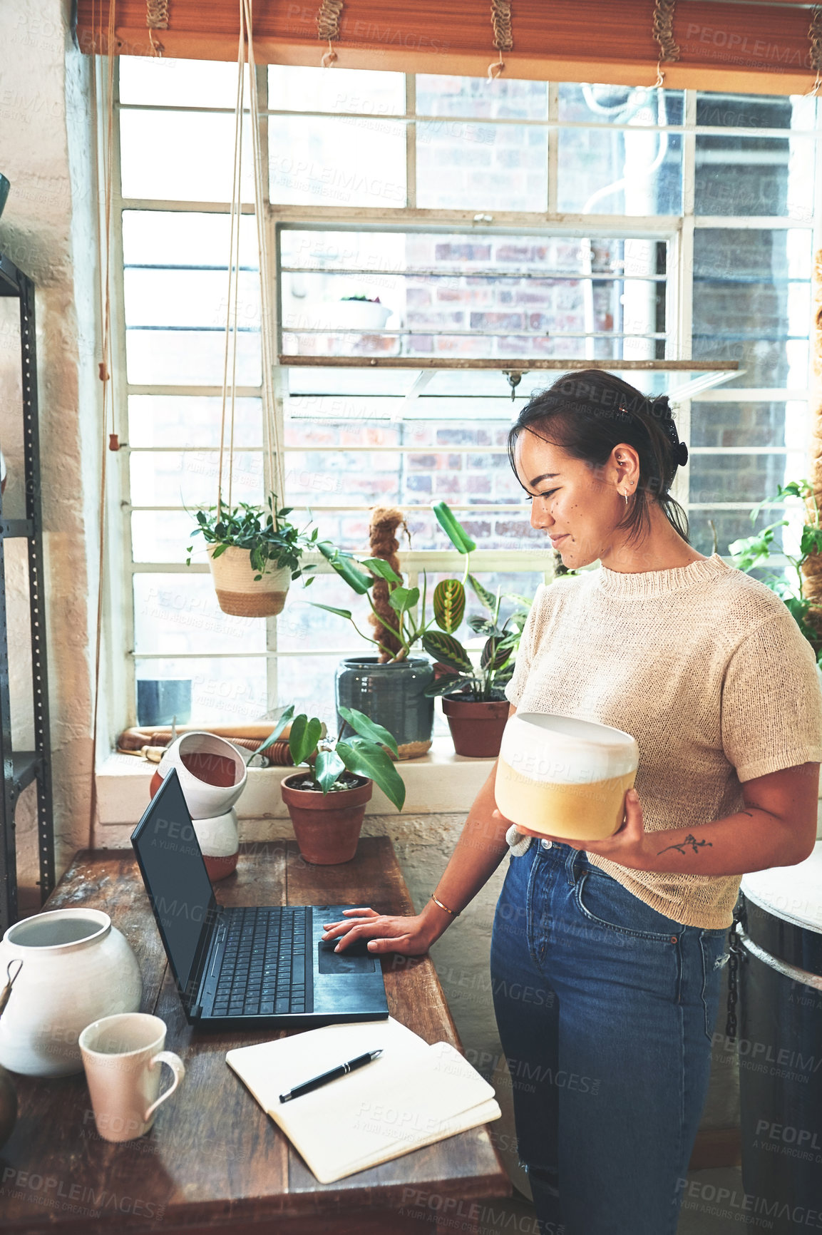 Buy stock photo Cropped shot of an attractive young business owner standing and holding a clay pot while using her laptop