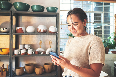 Buy stock photo Cropped shot of an attractive young business owner standing alone in her pottery studio and using her cellphone