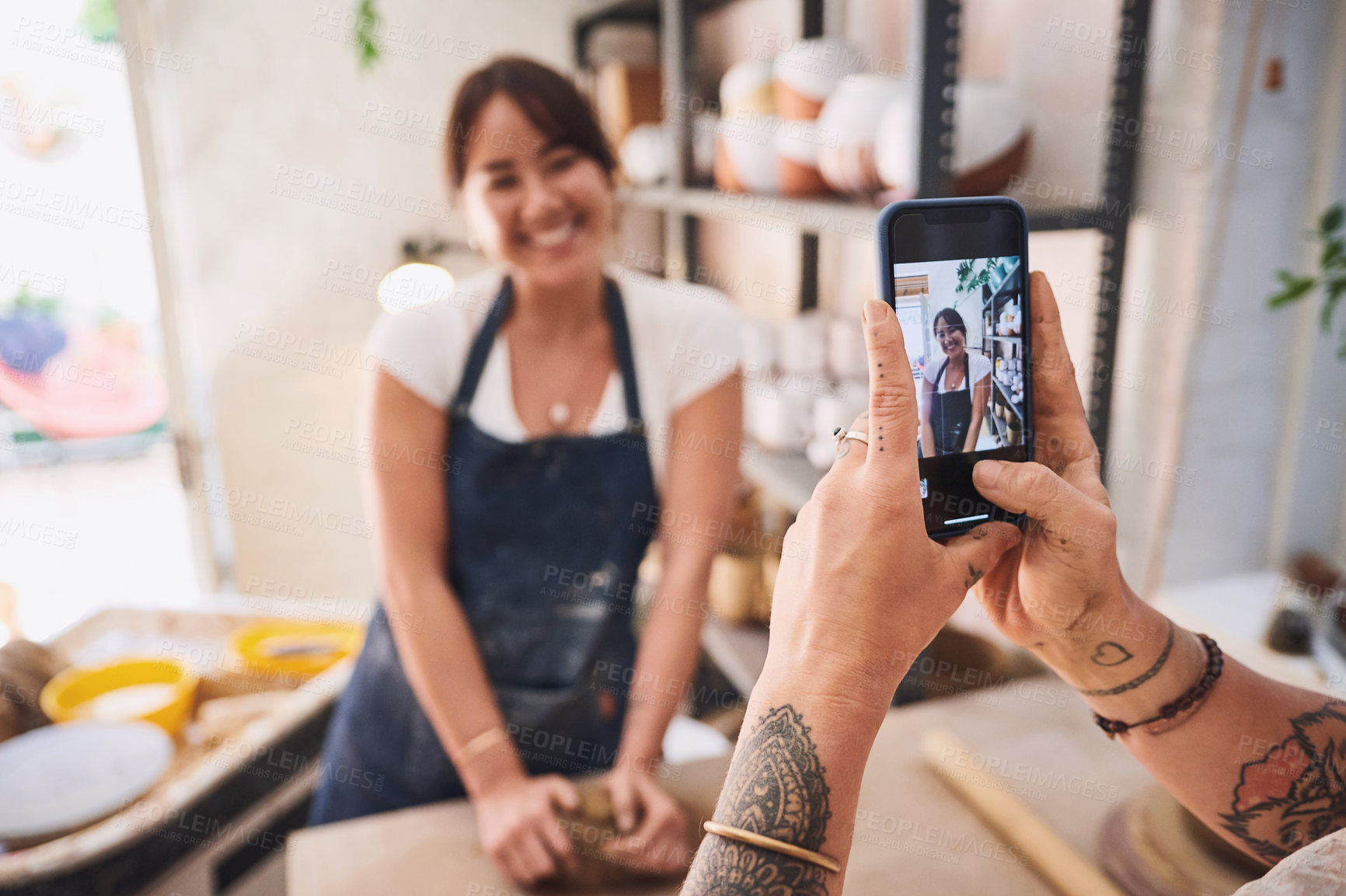 Buy stock photo Shot of a woman using a smartphone to take pictures of her friend in a pottery studio