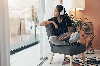 Buy stock photo Shot of a young woman using headphones with her smartphone and looking thoughtfully out of a window at home
