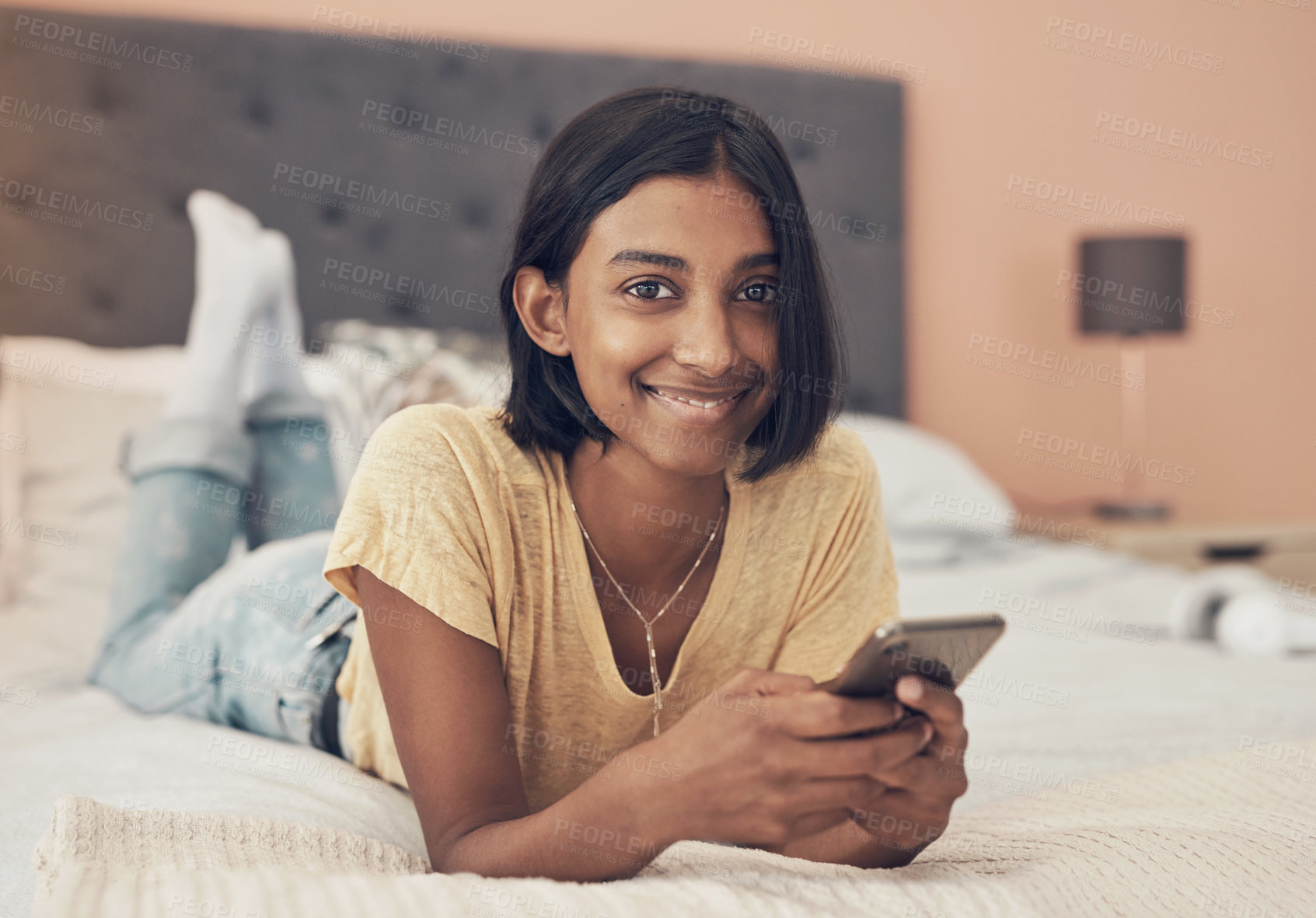 Buy stock photo Portrait of a young woman using a smartphone while relaxing on her bed at home