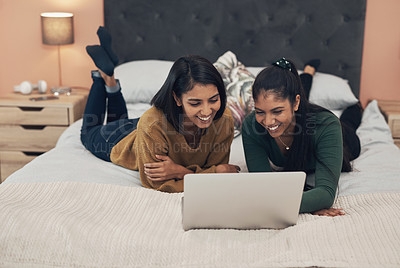 Buy stock photo Shot of two young women using a laptop while lying together on a bed