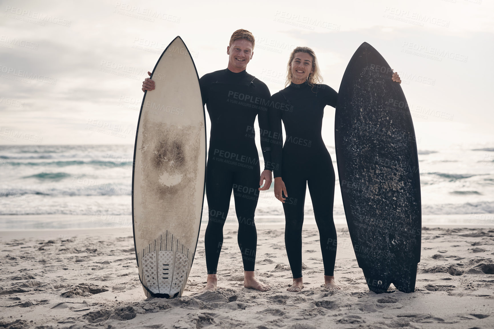 Buy stock photo Shot of a young couple standing on the beach with their surfboards