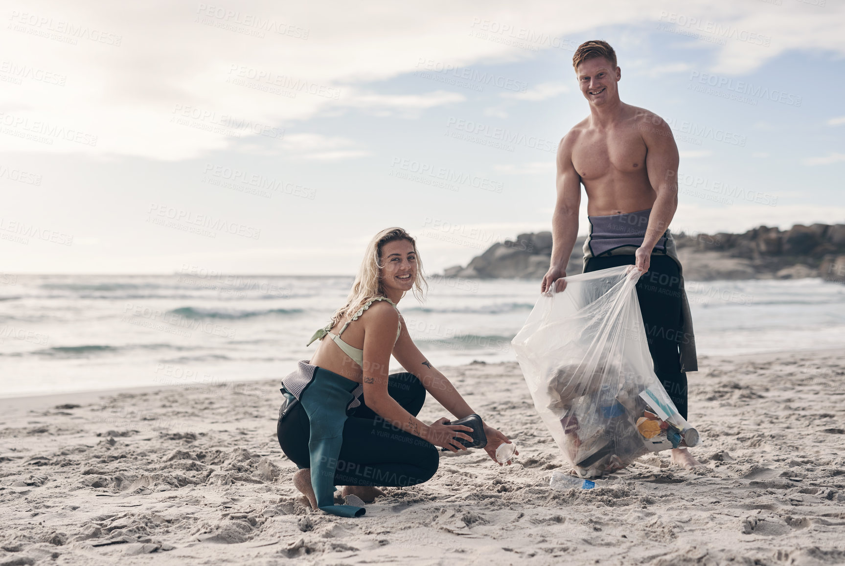 Buy stock photo Shot of a young couple doing a beach clean while wearing wetsuits