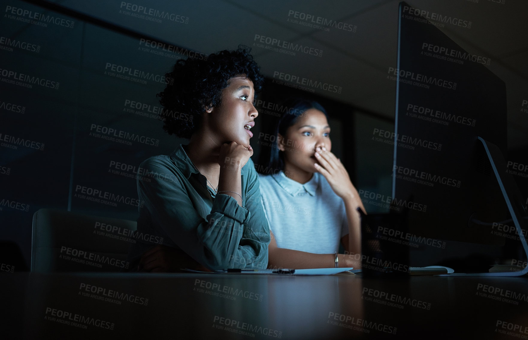 Buy stock photo Shot of two young businesswomen looking shocked while using a computer during a late night at work