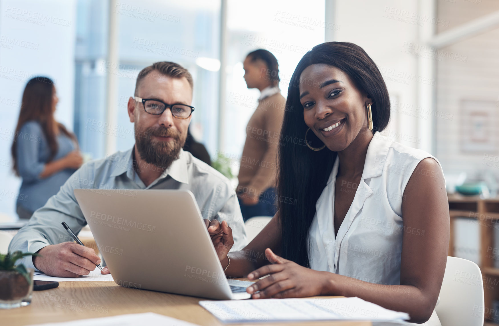 Buy stock photo Cropped portrait of two young businesspeople sitting together in the office while their colleagues work behind them