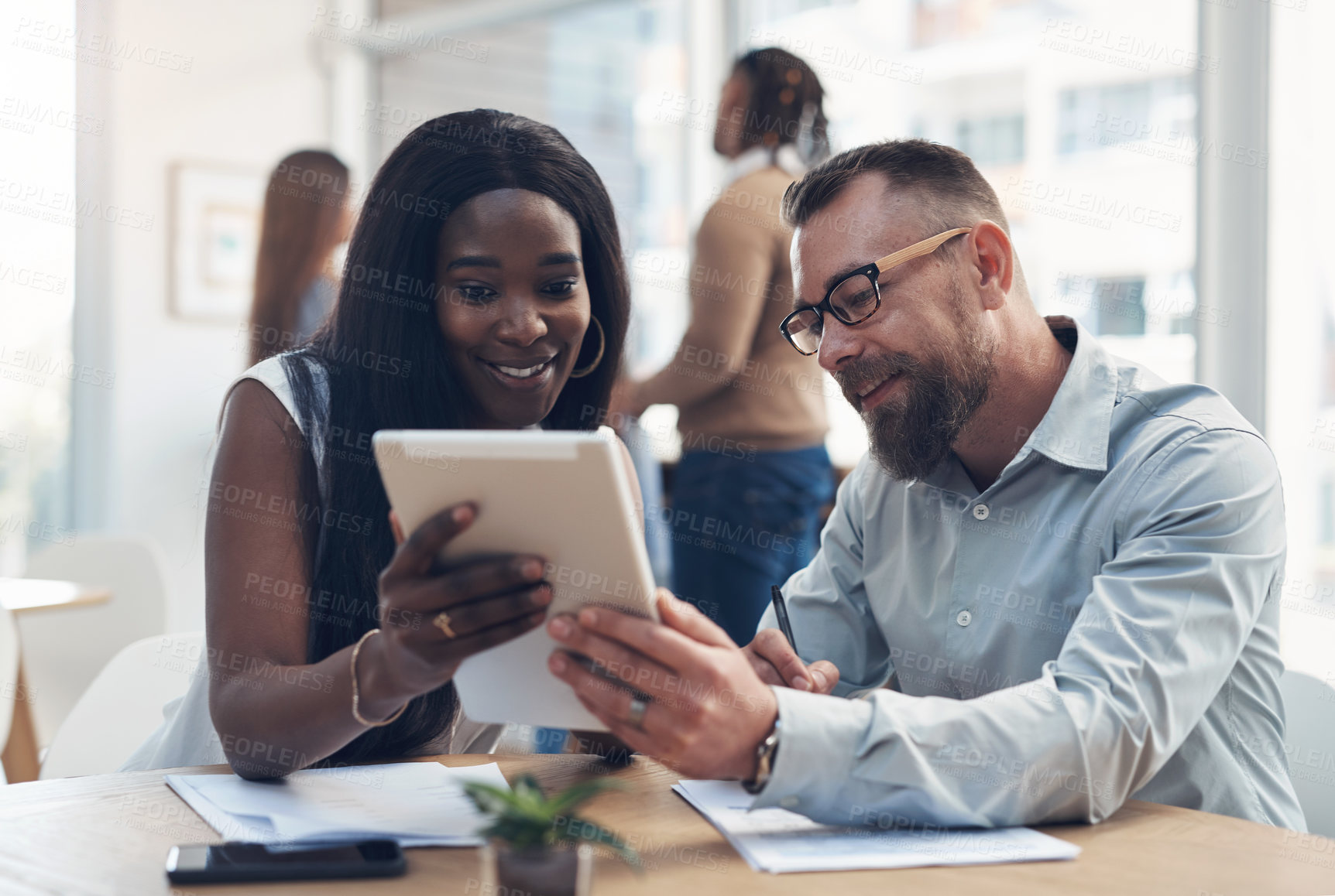 Buy stock photo Cropped shot of two young businesspeople sitting together and discussing in the office while their colleagues work behind them