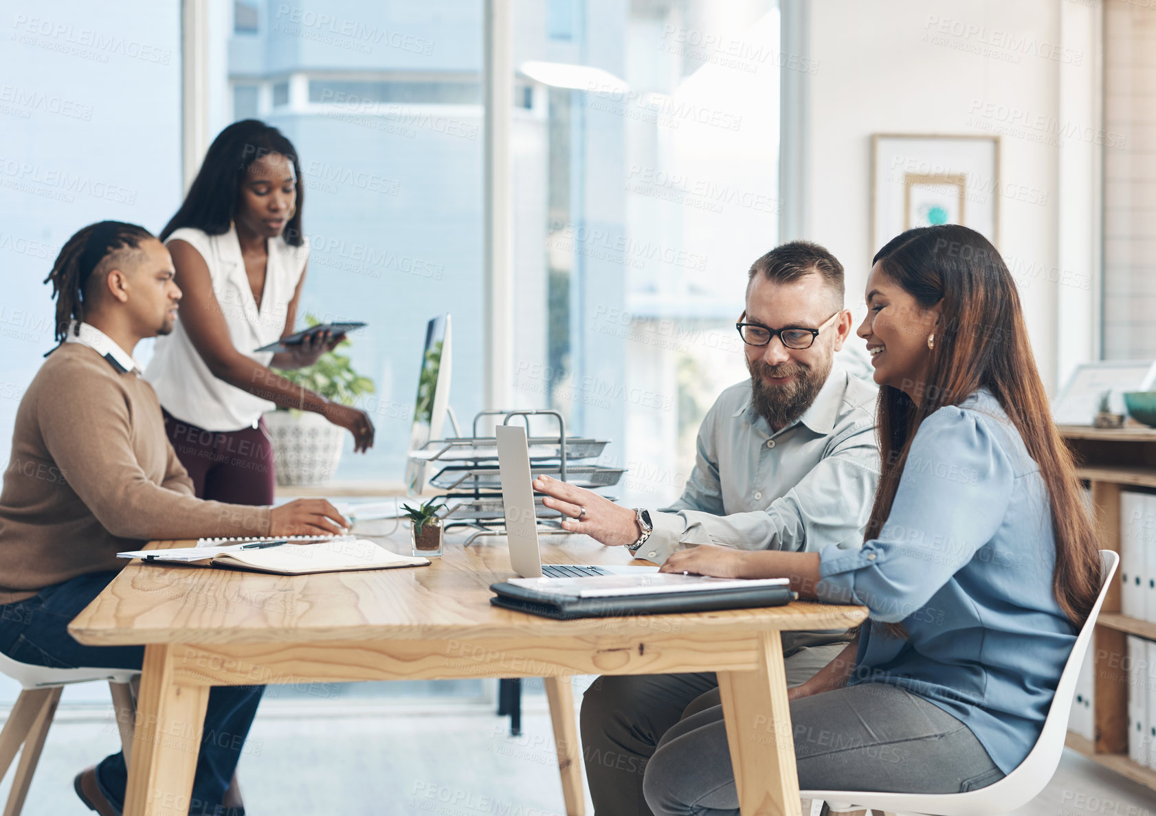 Buy stock photo Cropped shot of two young businesspeople sitting together and using a laptop while their colleagues work in the background