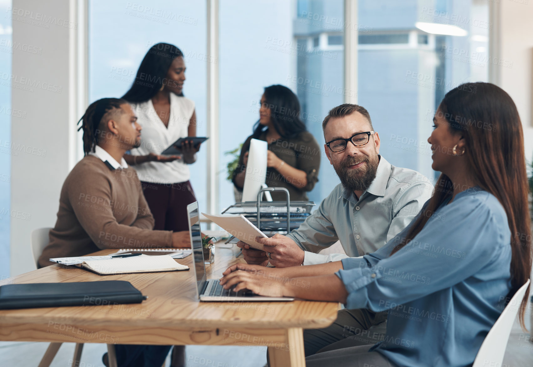 Buy stock photo Cropped shot of two young businesspeople sitting together and using a laptop while their colleagues work in the background