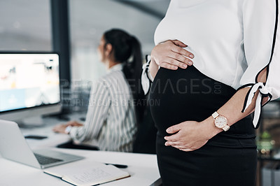 Buy stock photo Cropped shot of an unrecognizable pregnant businesswoman standing and holding her stomach while a colleague works behind her