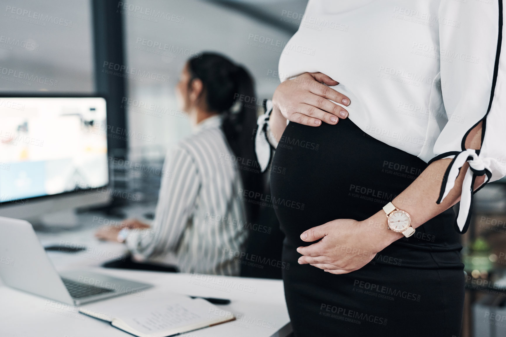 Buy stock photo Cropped shot of an unrecognizable pregnant businesswoman standing and holding her stomach while a colleague works behind her