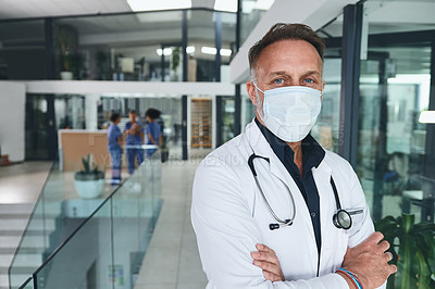 Buy stock photo Cropped portrait of a handsome mature doctor wearing a face mask and standing alone in the clinic