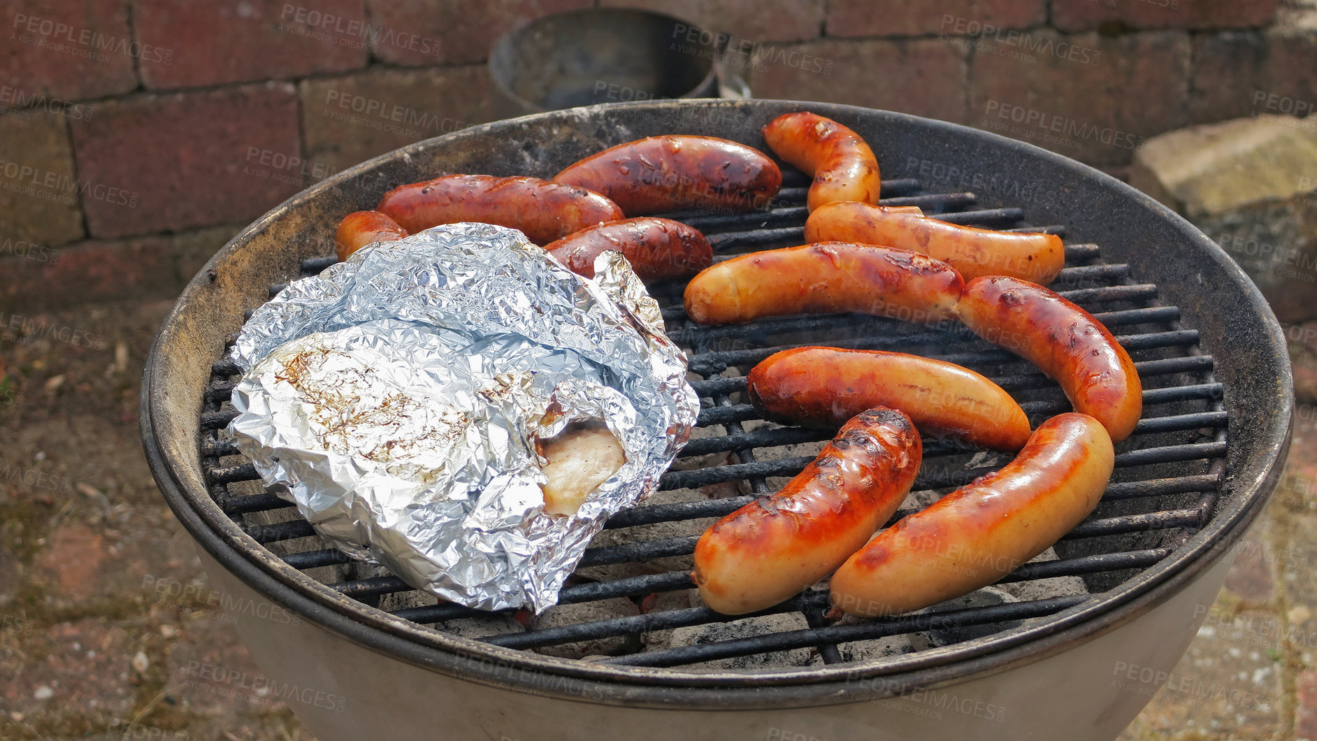 Buy stock photo Sizzling barbecue sausages and chicken wrapped in foil outdoors in a yard on a hot day. Delicious meat roast on a grill for a cookout by a home or house hosting a weekend party