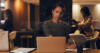 Buy stock photo Shot of a young businesswoman writing notes while working on a laptop in an office at night